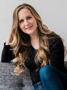 Headshot of woman sitting on the couch leaning on her arm. She has long, wavy light brown hair and is smiling