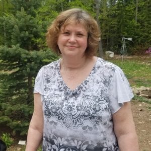 woman with short, dirt blonde curly hair smiling with her mouth closed in front of pine and green trees. She's wearing a gray and white short-sleeve shirt with designs on them.