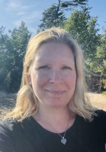 Woman with medium blond hair standing outside smiling with her mouth closed.