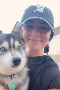 Woman holding a gray and white husky puppy, woman wearing a Minnesota jean hat and wearing black. Hair pulled back.