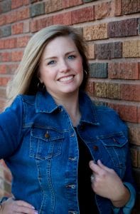 woman smiling with long blond hair leaning against a brick wall wearing a jean jacket.