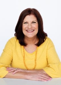 woman with medium brown hair smiling, wearing a bright yellow blouse and a long yellow beaded necklace
