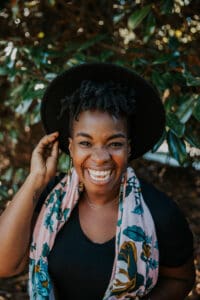 Headshot of a woman with dark black hair smiling huge. She's wearing a hat that one hand is touching, a pink flower scarf, and there is a tree in the background