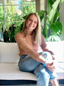 Woman with medium blonde hair sitting on a white couch with a window and greenery in the background. She has her legs crossed and her arms clasped in front of them. She is smiling